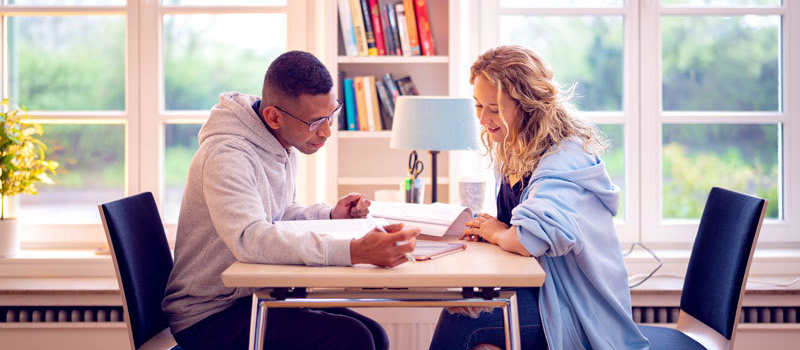 Zwei Studierende (Mann und Frau) sitzen gegenüber an einem Schreibtisch und schauen Lehrmaterialien durch.