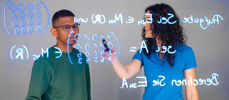 Studierende (Mann und Frau) stehen vor einem Lighboard. Darauf ist eine mathematische Formel geschrieben.