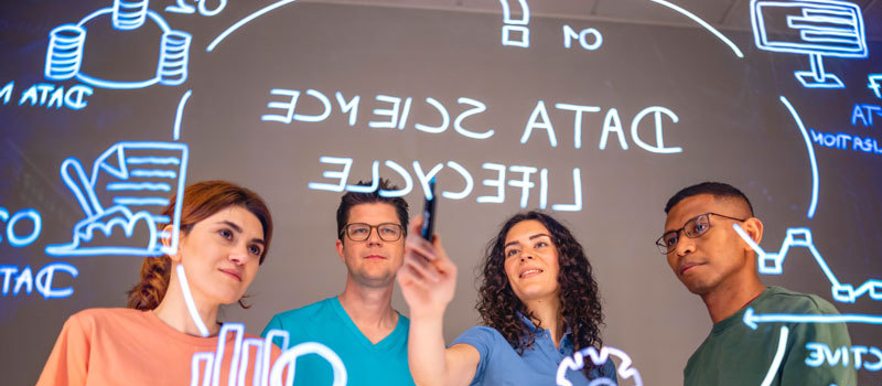 Eine Gruppe von Menschen steht vor einem Lightboard. Zu sehen ist der Data Science-Lifecycle, der dort aufgemalt ist. 