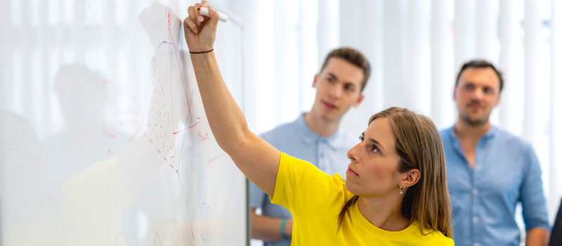 Eine Frau steht an einem Whiteboard. Zwei Studierende sind im Hintergrund.