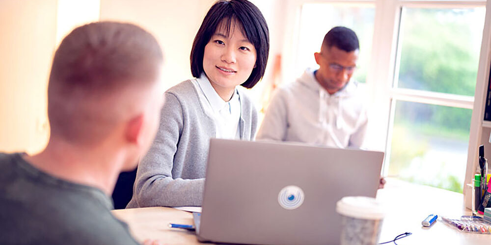 Woman sitting at a table with an open laptop looking at a man, another man in a hoodie sitting by a window in the background