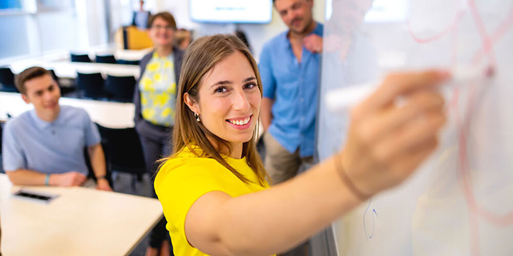 Junge Frau in gelbem T-Shirt schreibt an ein Whiteboard, im Hintergrund vier Personen in einem Klassenraum.