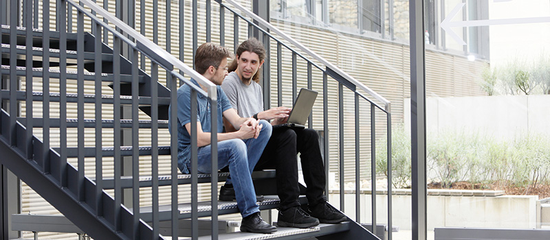 Zwei junge Männer sitzen mit Laptop auf Treppenstufen