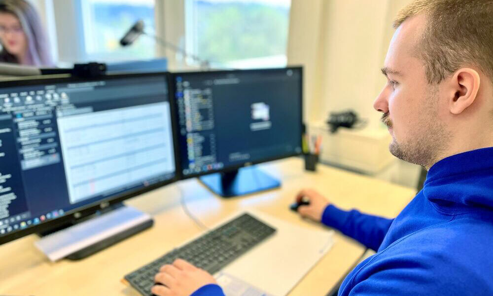 Junger Mann in blauem Pullover arbeitet an einem Schreibtisch mit zwei Monitoren und Tastatur in einem Büro