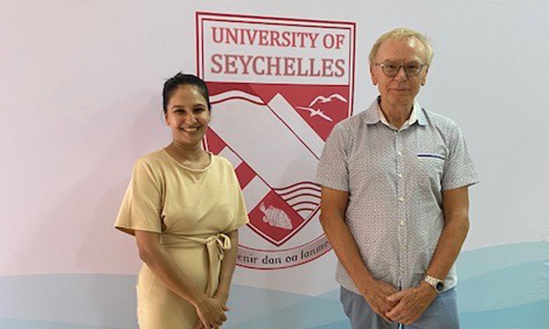 Foto von Prof. Helmut Wagner und einer weiteren weiblichen Person mit dunkler Hautfarbe vor einer Wand mit einem Emblem 