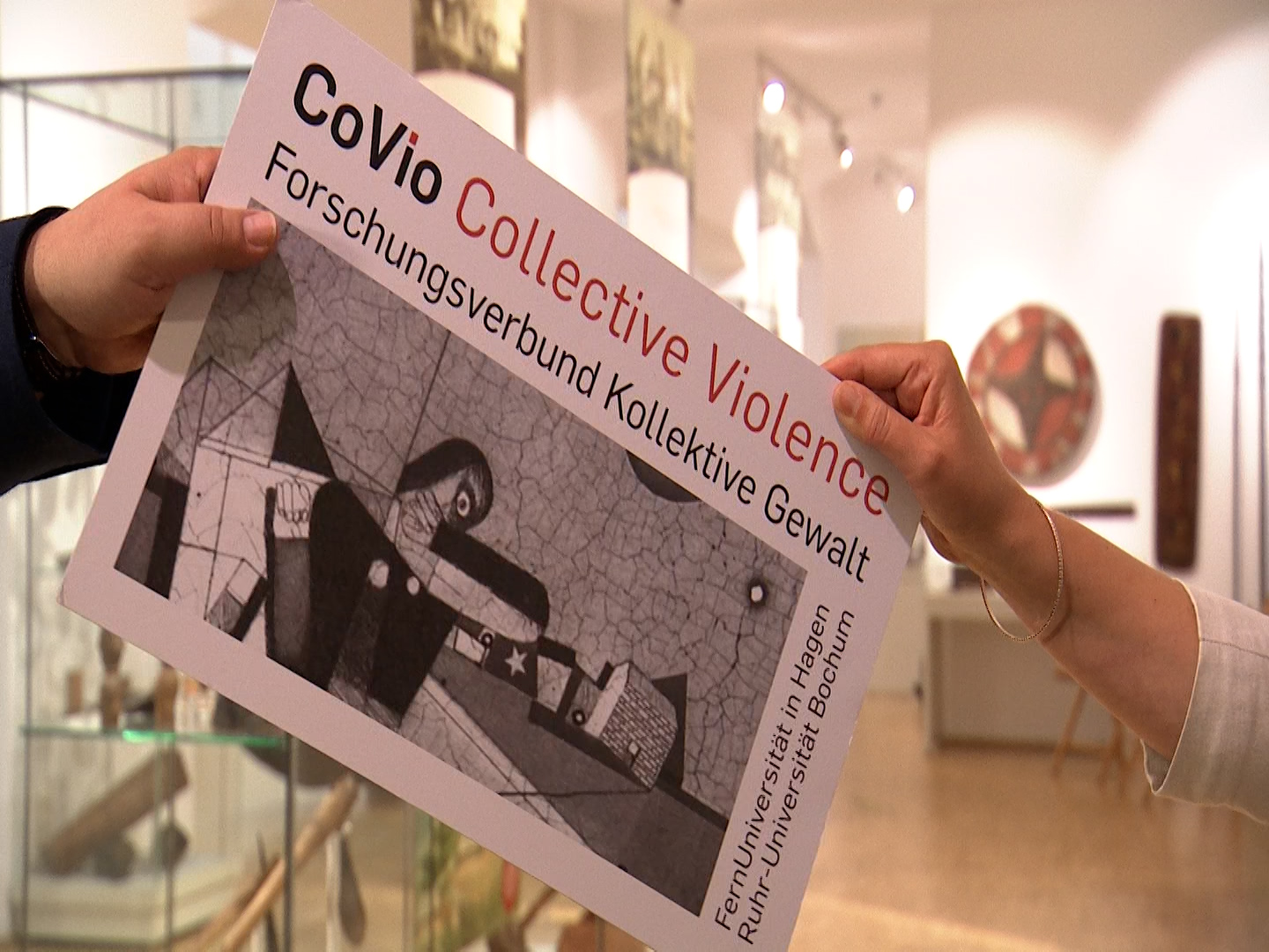 Zwei Hände reichen sich in einem Ausstellungsraum eine Pappe mit dem Logo des Forschungsverbundes CoVio Collective Violence