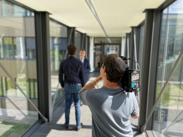 Eine Person mit Schulterkamera filmt zwei weitere, die sich in einem langen Flur unterhalten