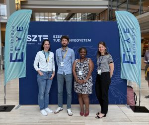 Foto von Réka Futász, Dr André Biedebeck, Dr Prue Goredema und Lídia Vinczéné Fekete vor einem Display der Tagung "European First Year Experience"